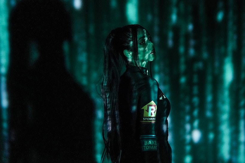 A person stands in profile against a backdrop of cascading green digital code, reminiscent of a futuristic or cyberpunk aesthetic. The person is wearing dark clothing with visible logos and has long hair. Shadows and green light interplay across their face and body, creating a mysterious atmosphere.
