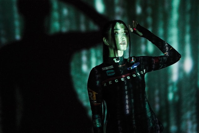 A person stands against a backdrop of projected green and black digital patterns, creating a futuristic atmosphere. The clothing features text such as 'RECESSION' and various logos. Shadows enhance the dramatic and immersive effect.