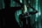 A person stands against a backdrop of projected green and black digital patterns, creating a futuristic atmosphere. The clothing features text such as 'RECESSION' and various logos. Shadows enhance the dramatic and immersive effect.