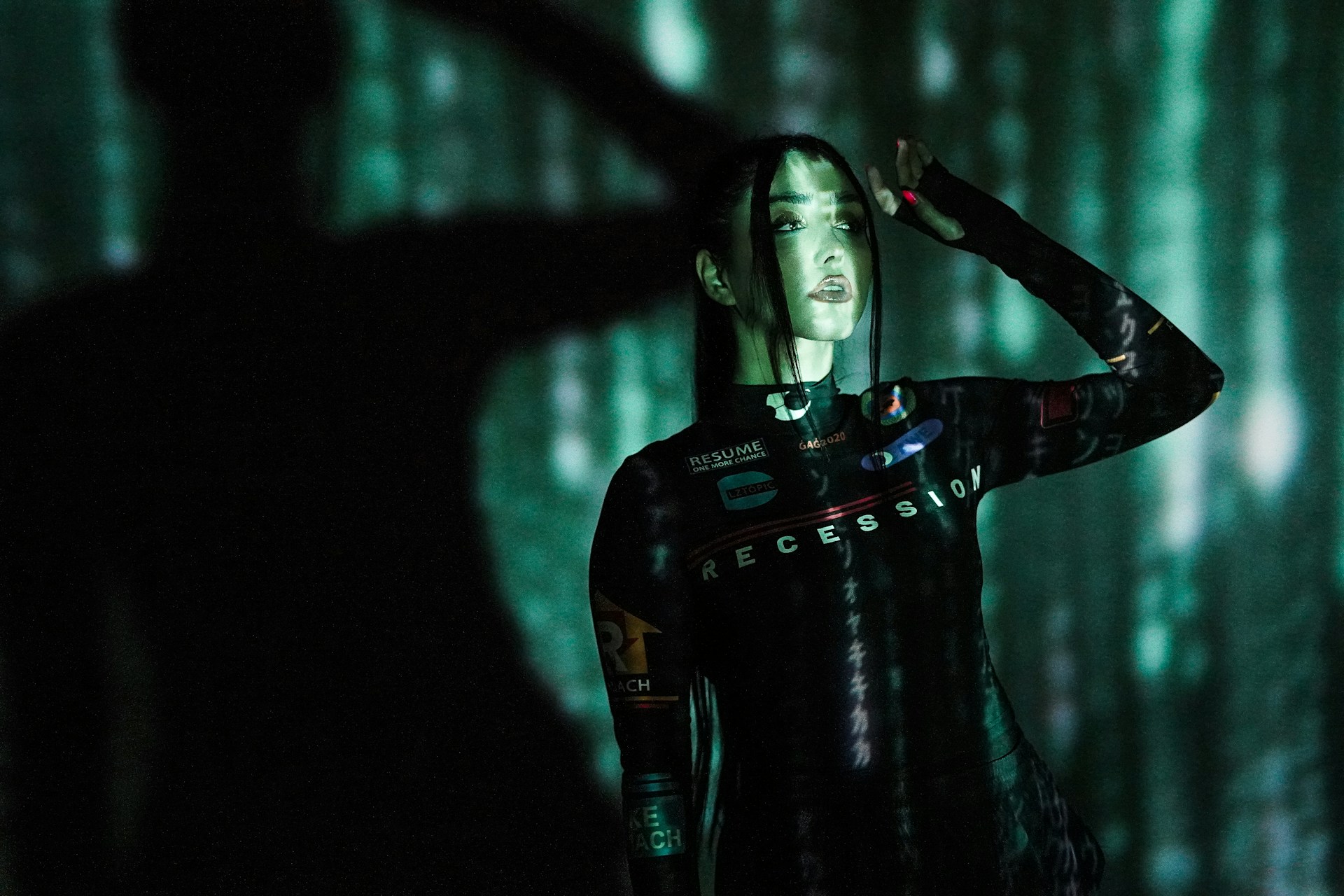 A person stands against a backdrop of projected green and black digital patterns, creating a futuristic atmosphere. The clothing features text such as 'RECESSION' and various logos. Shadows enhance the dramatic and immersive effect.