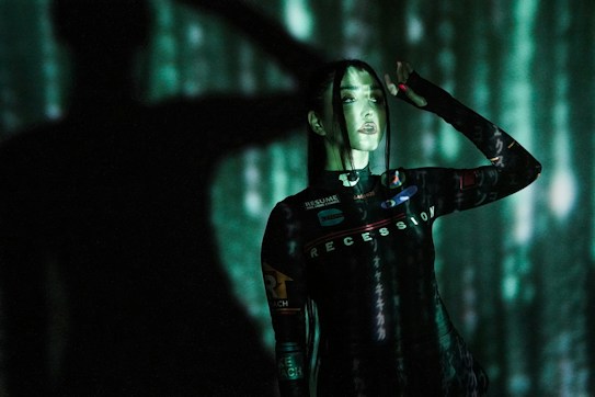 A person stands against a backdrop of projected green and black digital patterns, creating a futuristic atmosphere. The clothing features text such as 'RECESSION' and various logos. Shadows enhance the dramatic and immersive effect.