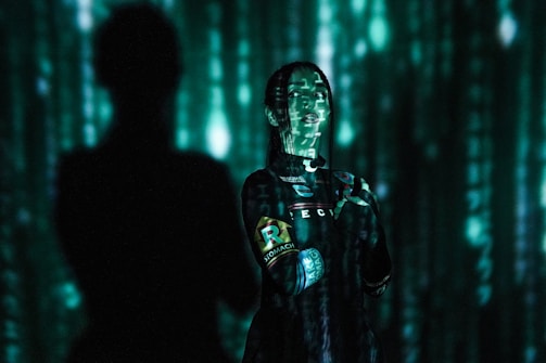 A person stands against a backdrop of green digital code patterns, reminiscent of a futuristic or virtual reality environment. The lighting creates dramatic shadows and highlights on the person's face and clothing.