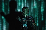 A person stands against a backdrop illuminated with green light patterns resembling digital rain, creating a futuristic and mysterious atmosphere. The person's attire is covered with various logos and text, adding complexity to the image. A shadow is cast prominently to the side, enhancing the dramatic effect.