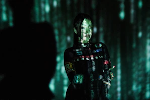 A person stands in a dark environment with green and blue digital patterns projected across their face and body. The patterns resemble falling digital code, creating a futuristic and mysterious ambiance. Their expression is calm, and they are wearing a dark outfit with various patches and logos.