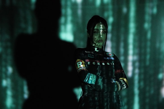 A person stands with arms crossed, partially enveloped in shadows with a digital light display projected on them, creating an atmosphere reminiscent of a digital or cyber setting.