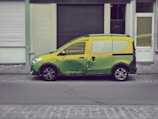 A small van painted in a blend of green and yellow hues is parked on a cobblestone street in front of a row of buildings with closed shutters.