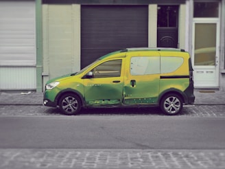 A small van painted in a blend of green and yellow hues is parked on a cobblestone street in front of a row of buildings with closed shutters.