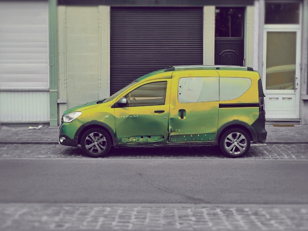 A small van painted in a blend of green and yellow hues is parked on a cobblestone street in front of a row of buildings with closed shutters.