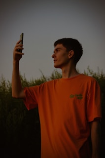 A person with short dark hair is holding a smartphone in front of them, possibly taking a photo or video in an outdoor setting at dusk. They are wearing an orange t-shirt and standing against a backdrop of greenery.