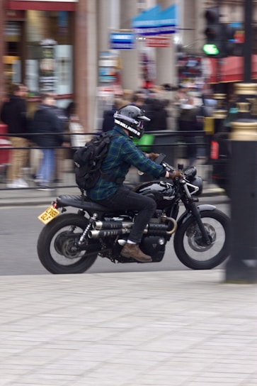 A person wearing a helmet and a backpack rides a motorcycle through an urban area. The bike is black with silver detailing. The background is slightly blurred, suggesting the bike is in motion, with several pedestrians and urban elements visible. Traffic lights and storefronts are part of the scene.