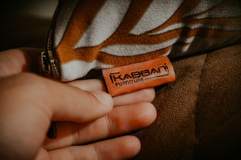 A hand is holding a brown leather tag with the text 'Kabbani Furniture' engraved on it. The tag is attached to a piece of upholstered furniture. The fabric of the furniture has a geometric pattern in shades of brown and white.
