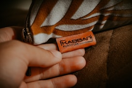 A hand is holding a brown leather tag with the text 'Kabbani Furniture' engraved on it. The tag is attached to a piece of upholstered furniture. The fabric of the furniture has a geometric pattern in shades of brown and white.
