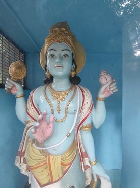 A statue of a deity with blue skin, adorned with golden ornaments, wearing a crown, and holding a conch shell and a disc. The figure is dressed in traditional attire with decorative patterns and has an assertive hand gesture. It is set against a blue background inside a room.