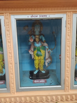 A statue of a deity enclosed in a glass case, holding a bow in one hand and arrows in a quiver on the back. The figure is adorned with a crown and traditional attire, featuring vibrant yellow and green garments. The surrounding case has beige ornamental carvings.