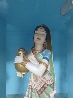 A statue of a woman in traditional attire holds a pot. Her attire includes a colorful saree with intricate patterns, adorned with jewelry such as earrings, necklaces, and bangles. The background is a light blue, complementing the vibrant colors of the statue.