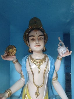 A detailed sculpture of a deity with intricate details, adorned with jewelry and holding a conch shell in one hand and a disc in the other. The figure is draped in a blue garment, set against a light blue background.