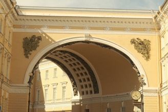 An ornate architectural arch connects two neoclassical buildings. Detailed sculptures and decorative elements are present on the arch's surface. A vintage clock with Roman numerals is mounted on a lamp post. The structures are painted in soft yellow tones, and the sky is partly cloudy.