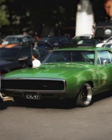 A vibrant green classic muscle car is parked among other vehicles, capturing attention with its sleek design. The scene suggests a car show or gathering, as there are multiple cars around and people in the background. The car is well-polished and features distinctive vintage styling.