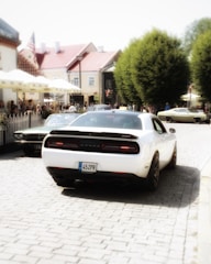 A white car with a license plate '45ZPR' is parked on a cobblestone street, flanked by other vintage cars. The scene is set in a quaint town with historic buildings and a crowd of people gathered nearby. Green trees line the edge of the street, adding a natural touch to the urban environment.