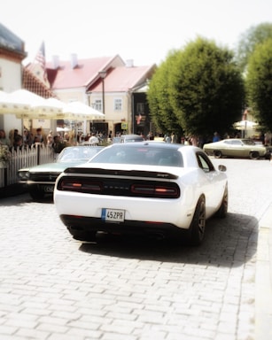 A white car with a license plate '45ZPR' is parked on a cobblestone street, flanked by other vintage cars. The scene is set in a quaint town with historic buildings and a crowd of people gathered nearby. Green trees line the edge of the street, adding a natural touch to the urban environment.