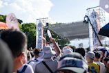A lively outdoor rally with supporters holding signs and cheering under a bright sky.