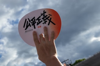 A hand holds up a fan against a backdrop of a cloudy sky. The fan is round with a red-orange gradient at the top and features bold black text along with a smaller line of text beneath it.