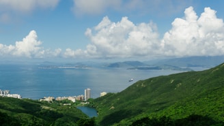 A scenic view of a coastline featuring expansive ocean waters, lush green hills, and scattered white clouds in a bright blue sky. Residential buildings are visible near the coastline, with a small body of water nestled among the greenery.