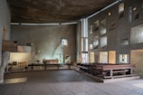 A spacious, minimally decorated interior of a chapel featuring rough-textured walls and modern architectural elements. The space includes wooden pews aligned on one side and a simple altar with candles and a cross. Natural light filters through small, irregularly shaped windows, casting subtle patterns on the walls.