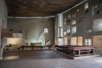 A spacious, minimally decorated interior of a chapel featuring rough-textured walls and modern architectural elements. The space includes wooden pews aligned on one side and a simple altar with candles and a cross. Natural light filters through small, irregularly shaped windows, casting subtle patterns on the walls.