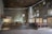 A spacious, minimally decorated interior of a chapel featuring rough-textured walls and modern architectural elements. The space includes wooden pews aligned on one side and a simple altar with candles and a cross. Natural light filters through small, irregularly shaped windows, casting subtle patterns on the walls.