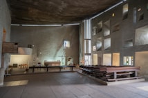A spacious, minimally decorated interior of a chapel featuring rough-textured walls and modern architectural elements. The space includes wooden pews aligned on one side and a simple altar with candles and a cross. Natural light filters through small, irregularly shaped windows, casting subtle patterns on the walls.