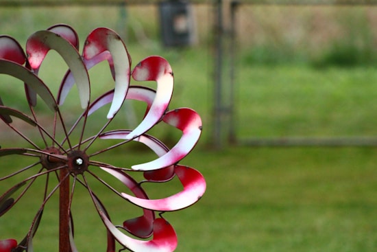 A metal garden wind spinner is depicted, featuring curving blades painted in a mix of red and white on a rusty metal structure. The background is blurred, showing green grass and a fence.