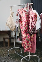A clothing rack is outdoors on a grassy area, displaying a variety of garments including a prominent floral dress with bright red and yellow flowers. A beige tote bag hangs from the left side of the rack with text printed on it. The surrounding environment includes a wooden bench and a metal structure.