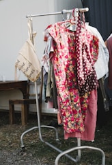 A clothing rack is outdoors on a grassy area, displaying a variety of garments including a prominent floral dress with bright red and yellow flowers. A beige tote bag hangs from the left side of the rack with text printed on it. The surrounding environment includes a wooden bench and a metal structure.