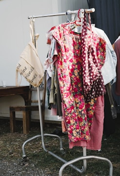 A clothing rack is outdoors on a grassy area, displaying a variety of garments including a prominent floral dress with bright red and yellow flowers. A beige tote bag hangs from the left side of the rack with text printed on it. The surrounding environment includes a wooden bench and a metal structure.