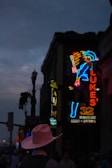 A neon-lit street scene at dusk with a cowboy-themed neon sign depicting a character in a blue outfit and hat. In front of the sign, a silhouette of a person wearing a pink cowboy hat is visible. The atmosphere is dim with streetlights and other illuminated signs visible in the background.