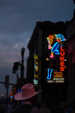 A neon-lit street scene at dusk with a cowboy-themed neon sign depicting a character in a blue outfit and hat. In front of the sign, a silhouette of a person wearing a pink cowboy hat is visible. The atmosphere is dim with streetlights and other illuminated signs visible in the background.