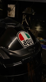 A close-up view of a black helmet prominently featuring the AGV logo with red, white, and green colors. The background is dimly lit, showing some blurred tools and equipment in a workshop setting.