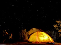 A glowing yellow tent is set up under a starry night sky, surrounded by sparse desert vegetation. A picnic table is partially visible to the left, indicating a campsite setting. The area is illuminated by the warm light coming from within the tent, creating a cozy atmosphere amidst the vast darkness of the night.