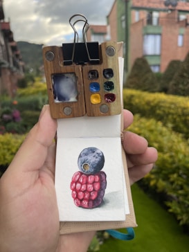 A small sketchbook is held in hand with a detailed watercolor painting of a blueberry and a raspberry. The sketchbook features a wooden cover with a clip holding the pages. A palette with various colors is attached to the top. In the background, there's an outdoor setting with greenery and buildings.