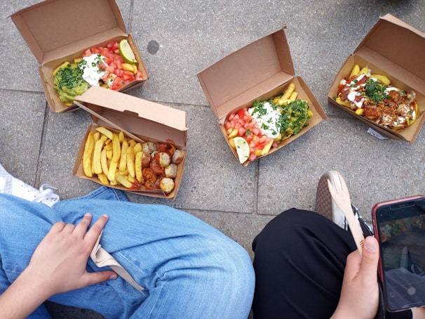 Several open cardboard boxes containing meals with fries, salad, sauces, and various toppings are placed on the ground. Two people are sitting nearby, each partially visible. One person is holding a wooden fork, and the other has a smartphone. The meals appear to be diverse in color and composition.