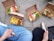 Several open cardboard boxes containing meals with fries, salad, sauces, and various toppings are placed on the ground. Two people are sitting nearby, each partially visible. One person is holding a wooden fork, and the other has a smartphone. The meals appear to be diverse in color and composition.