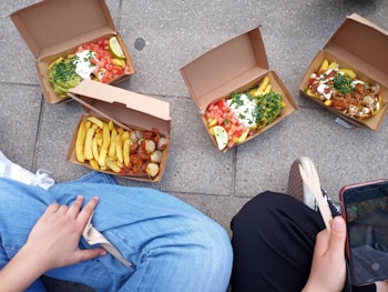 Several open cardboard boxes containing meals with fries, salad, sauces, and various toppings are placed on the ground. Two people are sitting nearby, each partially visible. One person is holding a wooden fork, and the other has a smartphone. The meals appear to be diverse in color and composition.