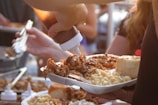 A person is holding a plate filled with macaroni and cheese, pulled pork, and a bread roll. Another hand is pouring barbeque sauce over the food from a squeeze bottle. The setting appears to be a communal or outdoor dining environment with other people nearby.
