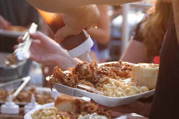 A person is holding a plate filled with macaroni and cheese, pulled pork, and a bread roll. Another hand is pouring barbeque sauce over the food from a squeeze bottle. The setting appears to be a communal or outdoor dining environment with other people nearby.