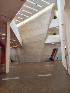 An interior view of a modern architectural space featuring large, angular concrete walls and a high ceiling with linear lighting. The floor is made of dark wood, adding contrast to the light-colored walls. In the distance, a person walks beside a table with chairs.