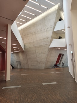 An interior view of a modern architectural space featuring large, angular concrete walls and a high ceiling with linear lighting. The floor is made of dark wood, adding contrast to the light-colored walls. In the distance, a person walks beside a table with chairs.
