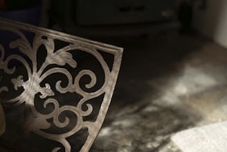 A decorative metal panel with intricate floral and scrollwork design is partially illuminated by soft light. It casts shadows on a textured surface beneath it, creating a play of light and shadow. The background is dimly lit, adding depth and contrast.