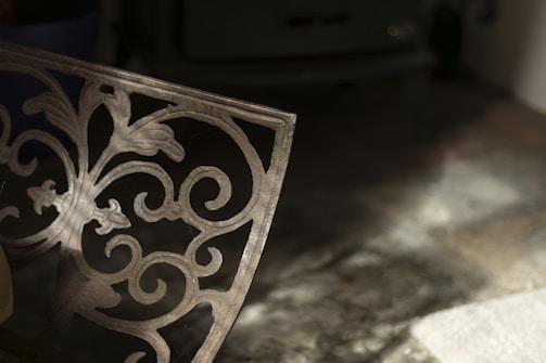 A decorative metal panel with intricate floral and scrollwork design is partially illuminated by soft light. It casts shadows on a textured surface beneath it, creating a play of light and shadow. The background is dimly lit, adding depth and contrast.