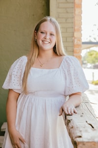 A young woman with long blonde hair is smiling while leaning against a rustic wooden surface. She is wearing a white dress with short, puffy sleeves. The background includes a green wall with brick accents and a hint of outdoor scenery visible through a window.